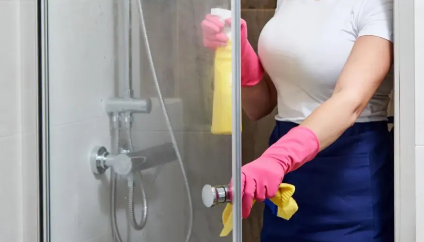 Persona con guantes rosas y delantal azul limpia la mampara del baño con spray amarillo.