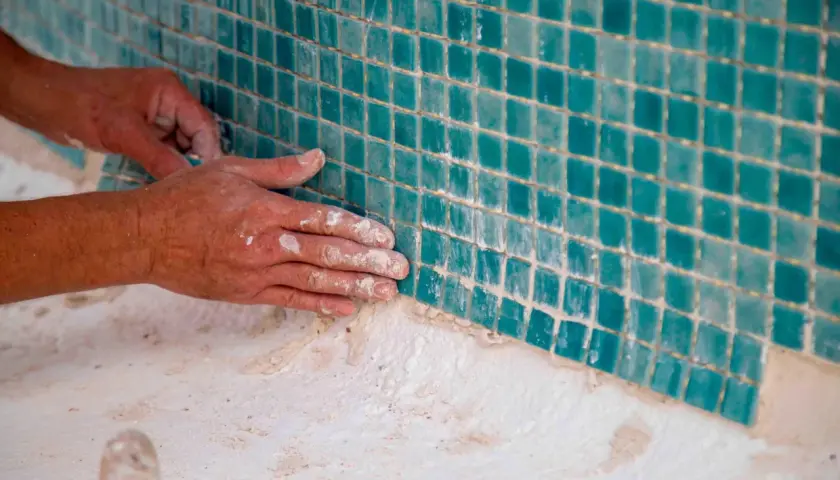 Manos trabajando en el cemento de piscinas elevadas de obra, colocando teselas turquesas.
