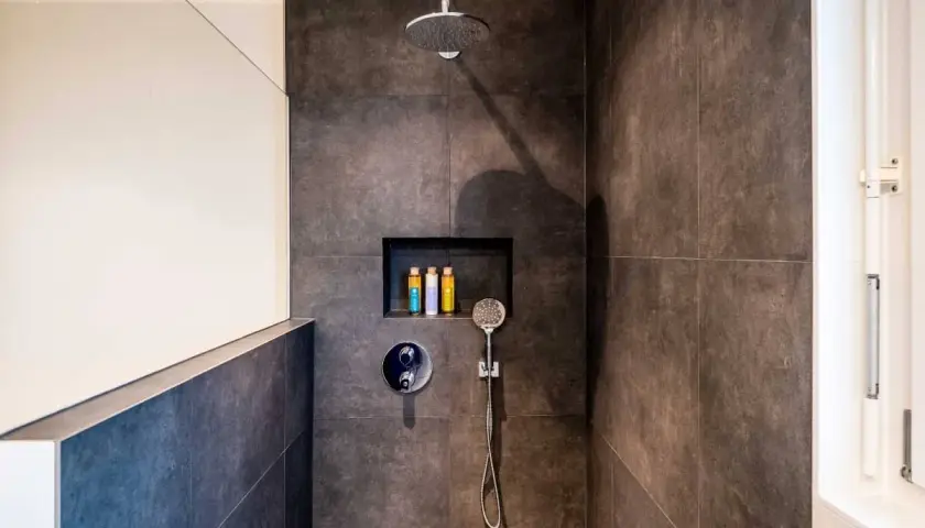 Duchas con murete y cristal. Cabezal de ducha, grifería y nicho con botes de champú. Azulejos oscuros y pared blanca.