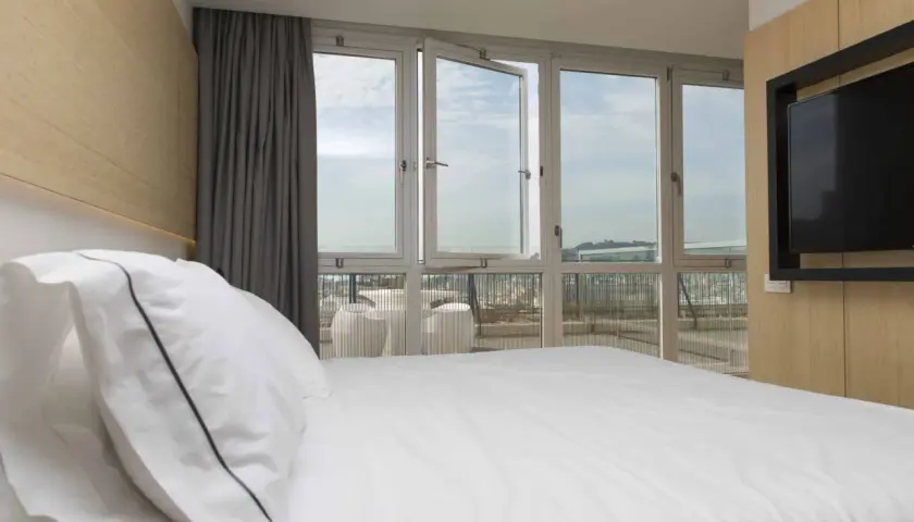 Cama blanca con almohadas, cortinas grises y ventanas de triple cristal que dejan ver el cielo y la ciudad.