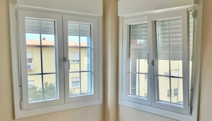 Dos ventanas batientes horizontales blancas con persianas enrollables. A través de ellas se ve un edificio amarillo con ventanas.
