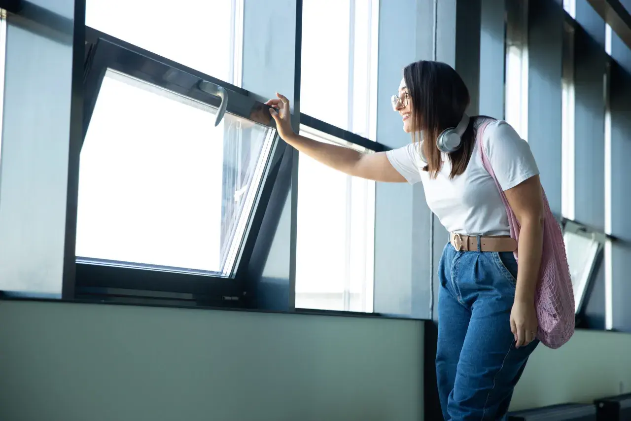 Mujer sonriente abre una ventana abatible, mostrando varios tipos de ventanas en un edificio moderno.
