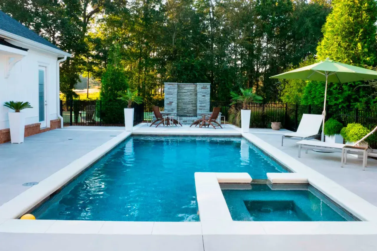 Piscina moderna con jacuzzi y cascada de piedra, lista para el mantenimiento de piscinas. Sillas, sombrilla y vegetación completan el oasis.