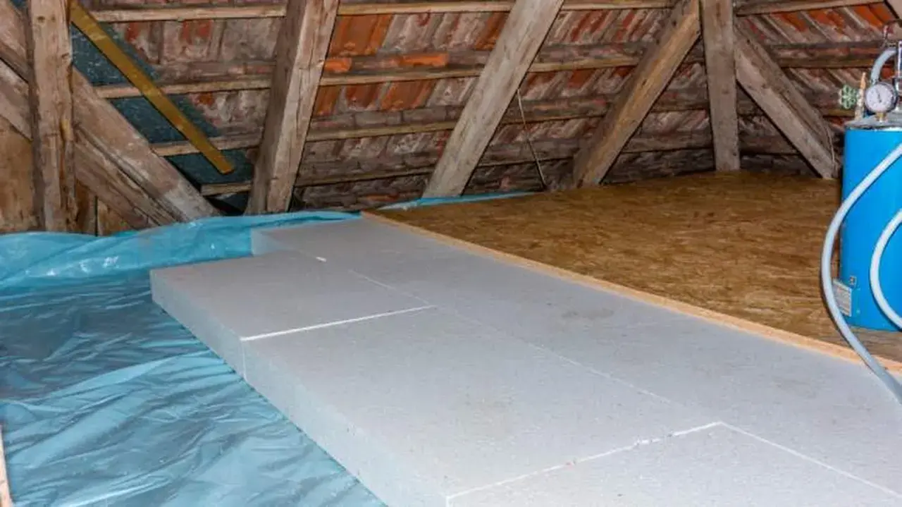 Aislamiento de suelo en una casa antigua con paneles de poliestireno sobre plástico azul, bajo vigas de madera y tejas.