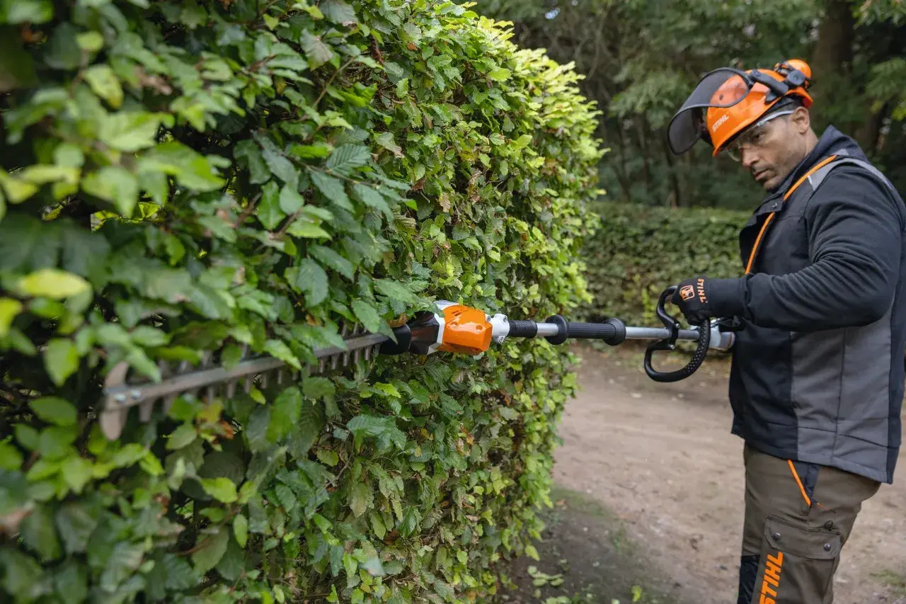 Hombre con equipo de protección realizando la poda de setos con una cortasetos eléctrica.