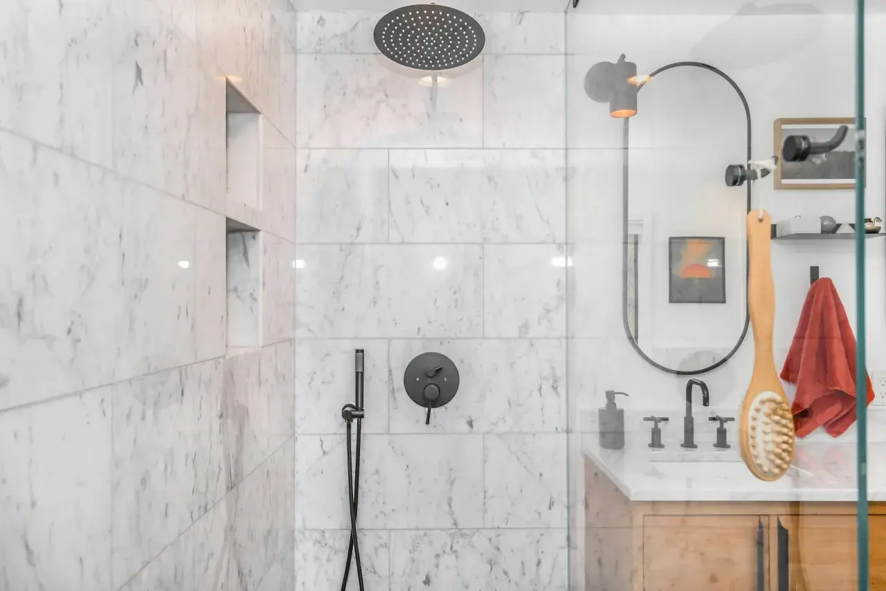 Ducha empotrada con grifo negro y rociador superior. La pared de mármol blanco tiene nichos empotrados.