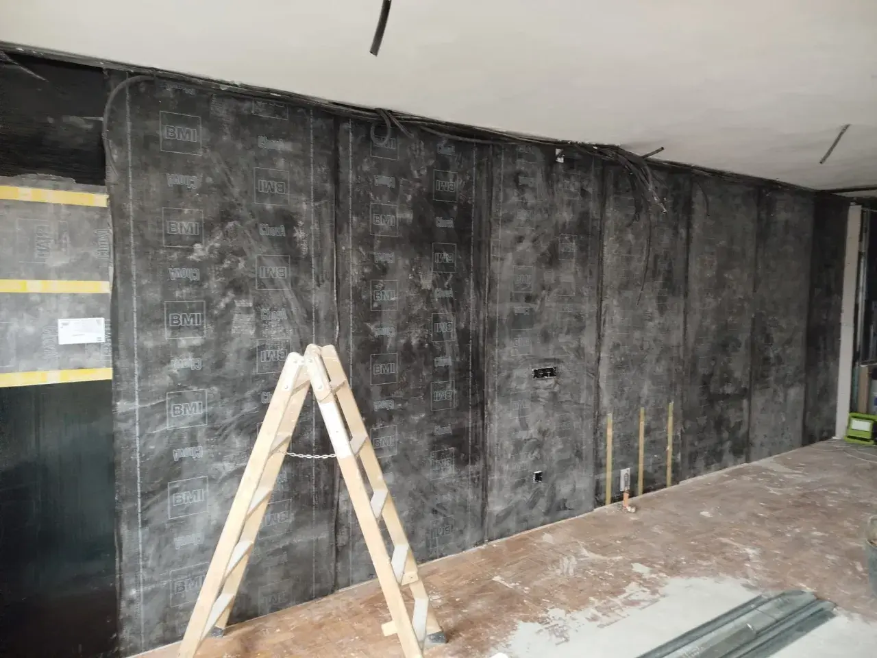 Instalación de material para insonorizar pared sin obra. Una escalera de madera apoya contra la pared cubierta con láminas negras.