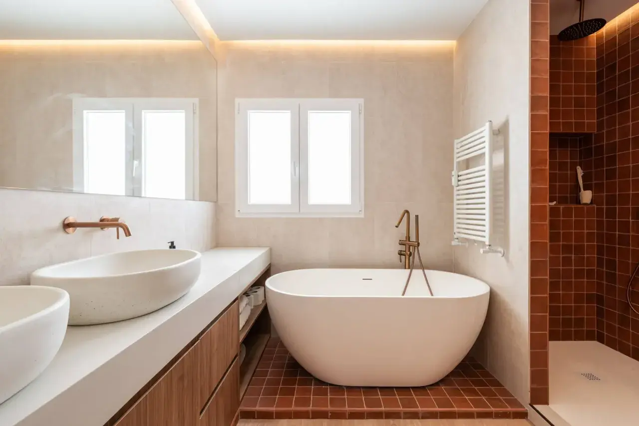 Baño moderno con bañera exenta y ducha. La iluminación indirecta en el techo y la luz natural de la ventana ayudan a como iluminar un baño.