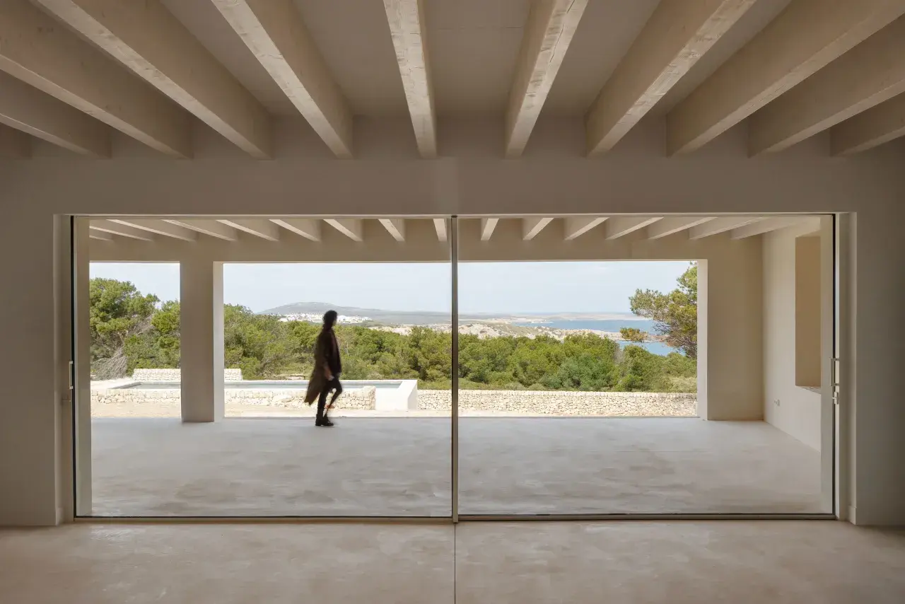 Persona camina por una terraza moderna con grandes ventanales que muestran varios tipos de ventanas correderas, vistas al mar y vegetación.
