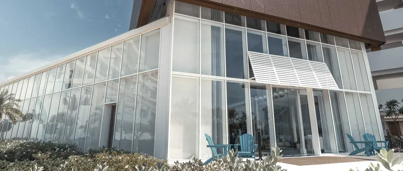 Edificio moderno con un **muro cortina fachada** de cristal. Dos sillas azules esperan en la entrada, reflejando palmeras.