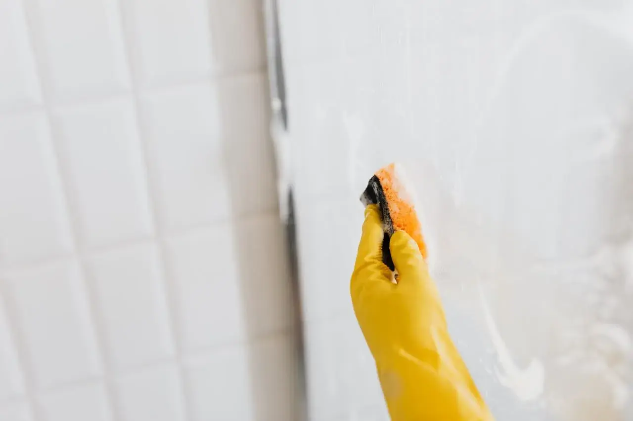 Mano con guante amarillo limpiando la mampara con esponja. Así se limpian las gomas de la mampara para un brillo impecable.