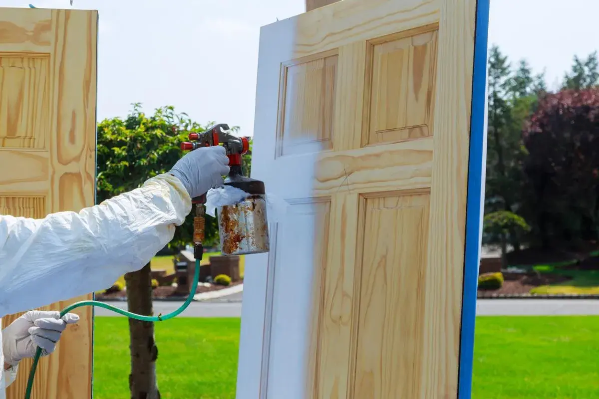 Persona con guantes y traje protector aplica pintura blanca a una puerta de madera con spray. Proceso de como lacar puertas barnizadas.