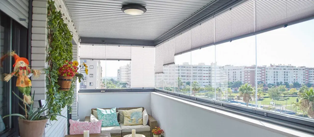 Balcón moderno con sofá, cojines coloridos y plantas. Las cortinas plisadas ofrecen privacidad, ideales para tus ideas para cubrir balcones.