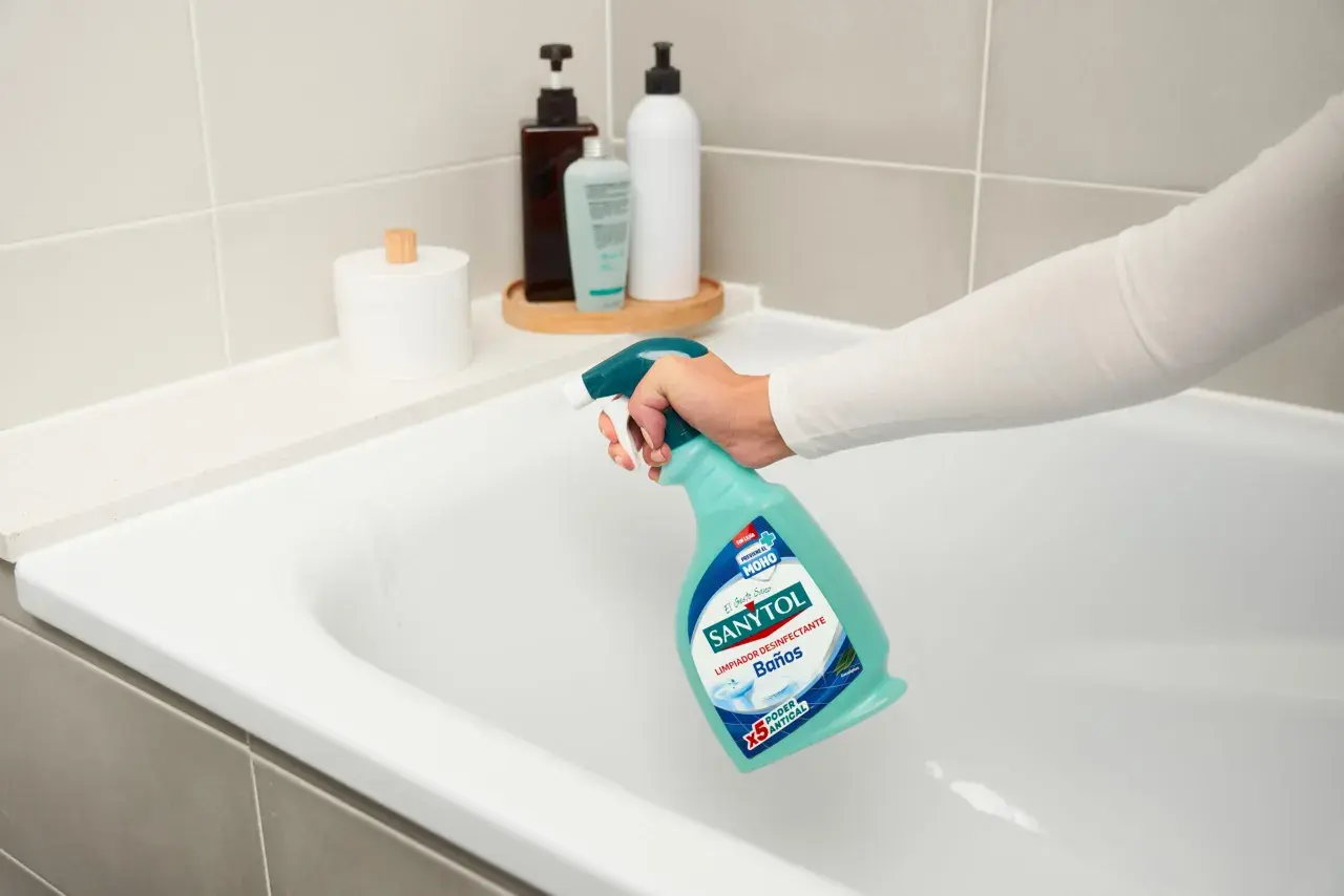 Mano rociando limpiador Sanytol en bañera blanca para quitar manchas oscuras plato ducha.