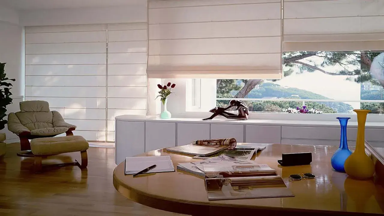Estudio moderno con sillón, mesa y varios tipos de persianas romanas que filtran la luz del sol.