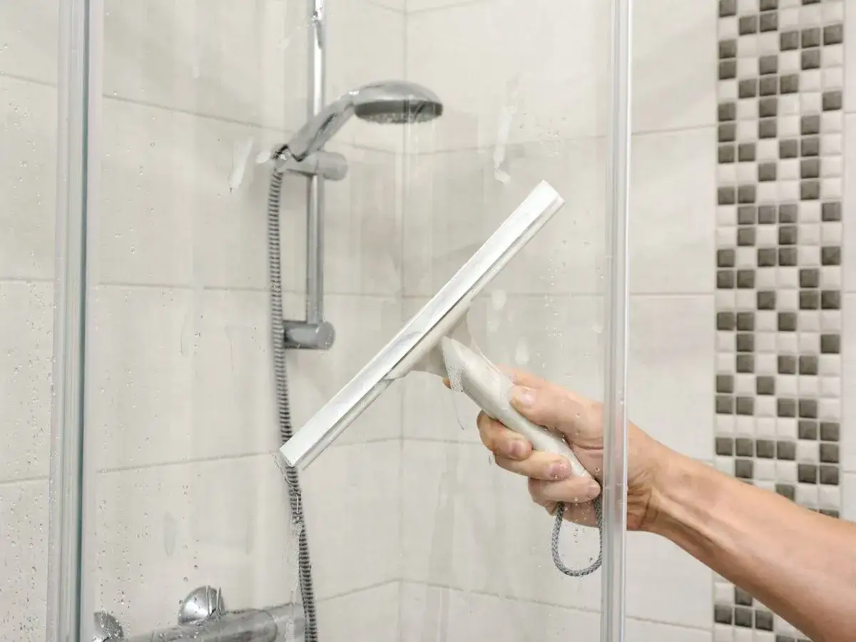 Mano limpiando la mampara del ba&ntilde;o con una rasqueta. Sigue estos pasos para saber como limpiar la mampara del ba&ntilde;o y dejarla reluciente.