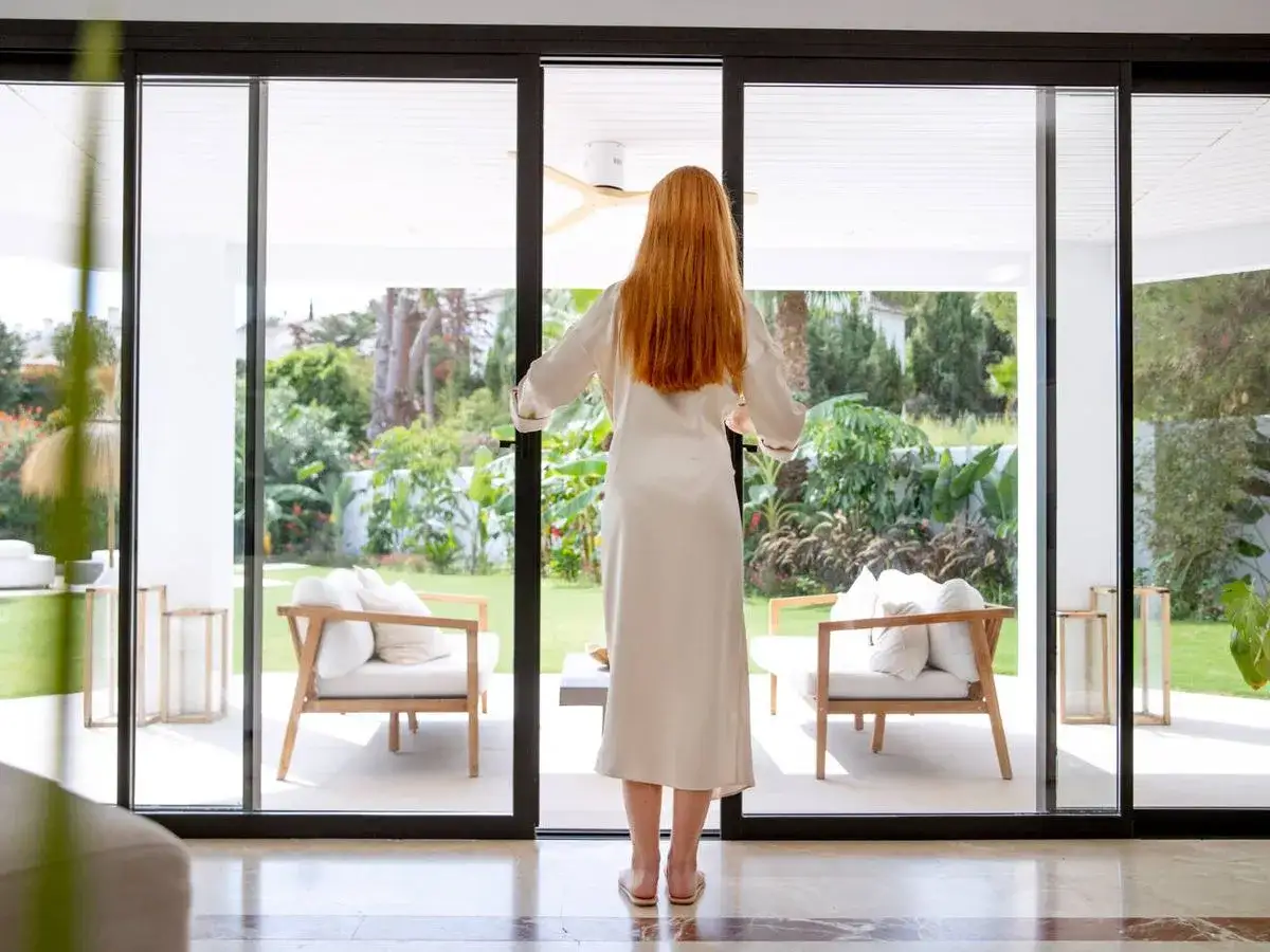 Mujer con cabello rojo abre puertas correderas de cristal, revelando un jard&iacute;n exuberante. La escena evoca la sensaci&oacute;n de sellar el espacio para disfrutar de la tranquilidad.