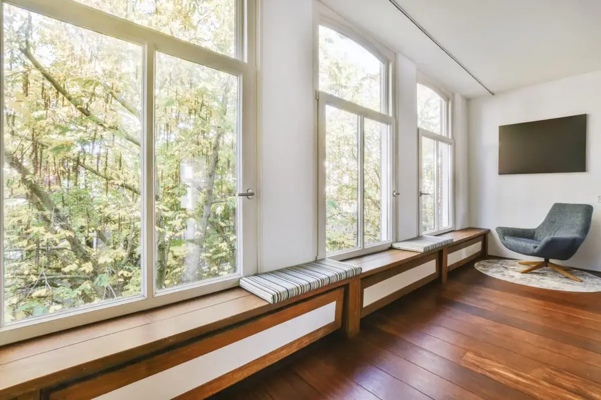 Amplia ventana de seguridad con bancos de madera y cojines, que ofrece vistas a &aacute;rboles frondosos.