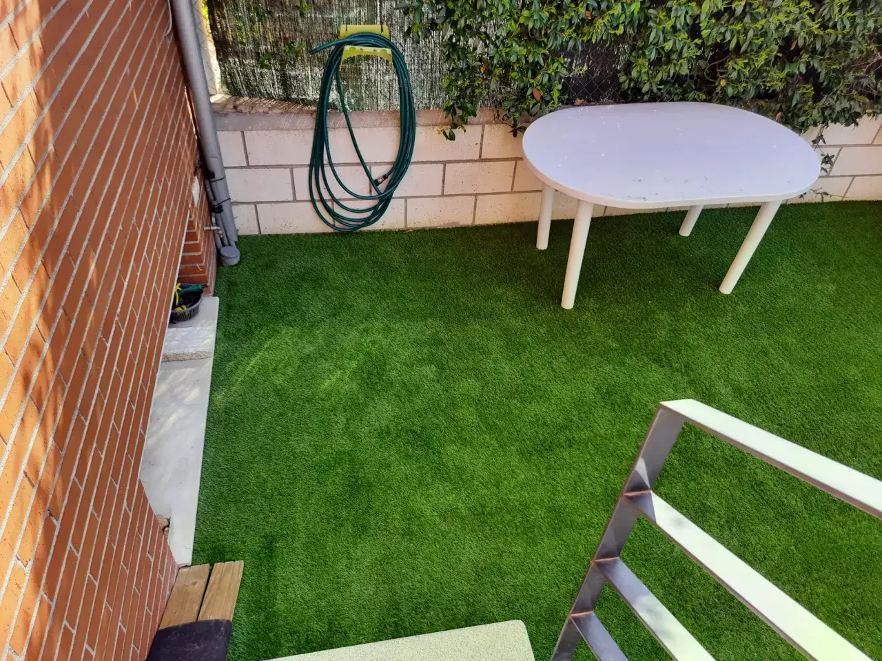 Un patio peque&ntilde;o con c&eacute;sped artificial, una mesa blanca y una manguera enrollada.