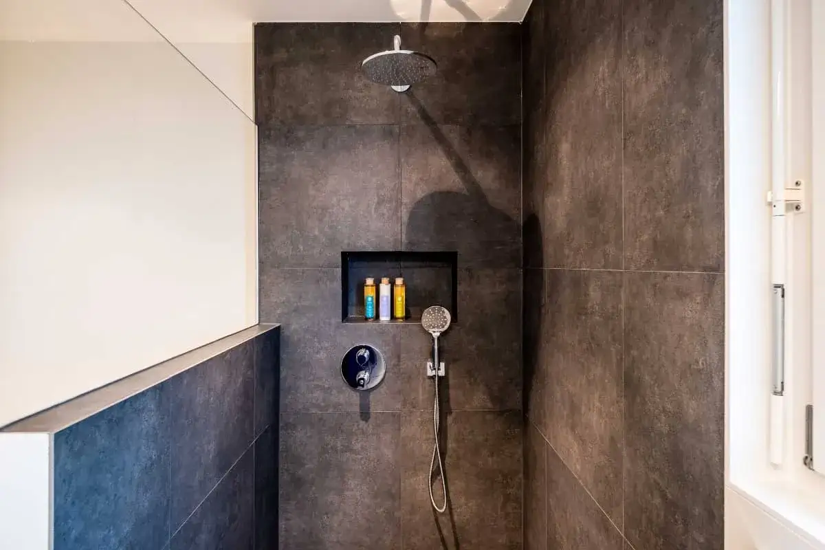 Ducha moderna con tabique de obra y muro de azulejos oscuros. Incluye nicho empotrado con botellas de champ&uacute; y ducha de mano.