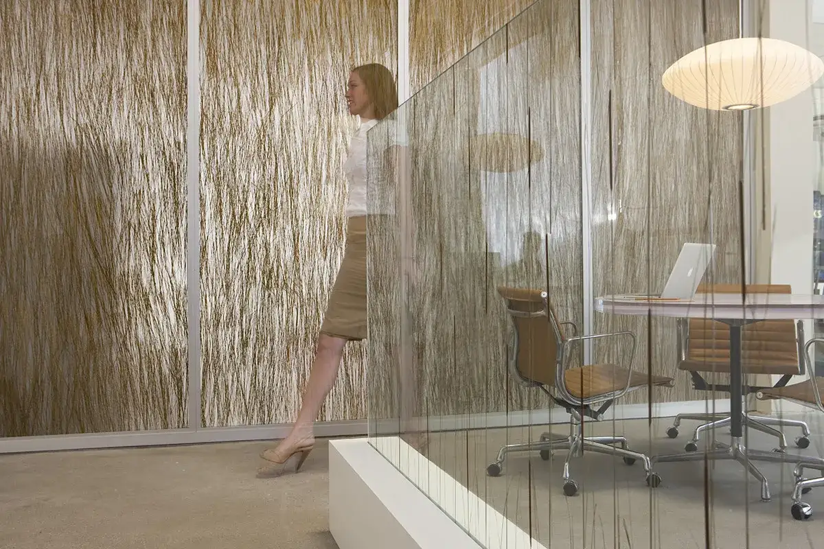 Una mujer camina junto a unas paredes que dejan pasar la luz, con un dise&ntilde;o que simula hebras secas. Al fondo, una sala de reuniones con sillas y mesa.
