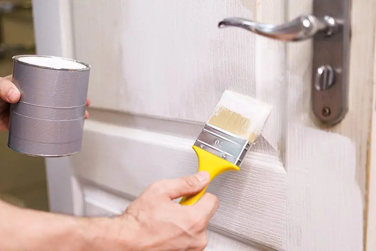 Persona aplica pintura blanca a una puerta de madera ya pintada con un pincel amarillo.
