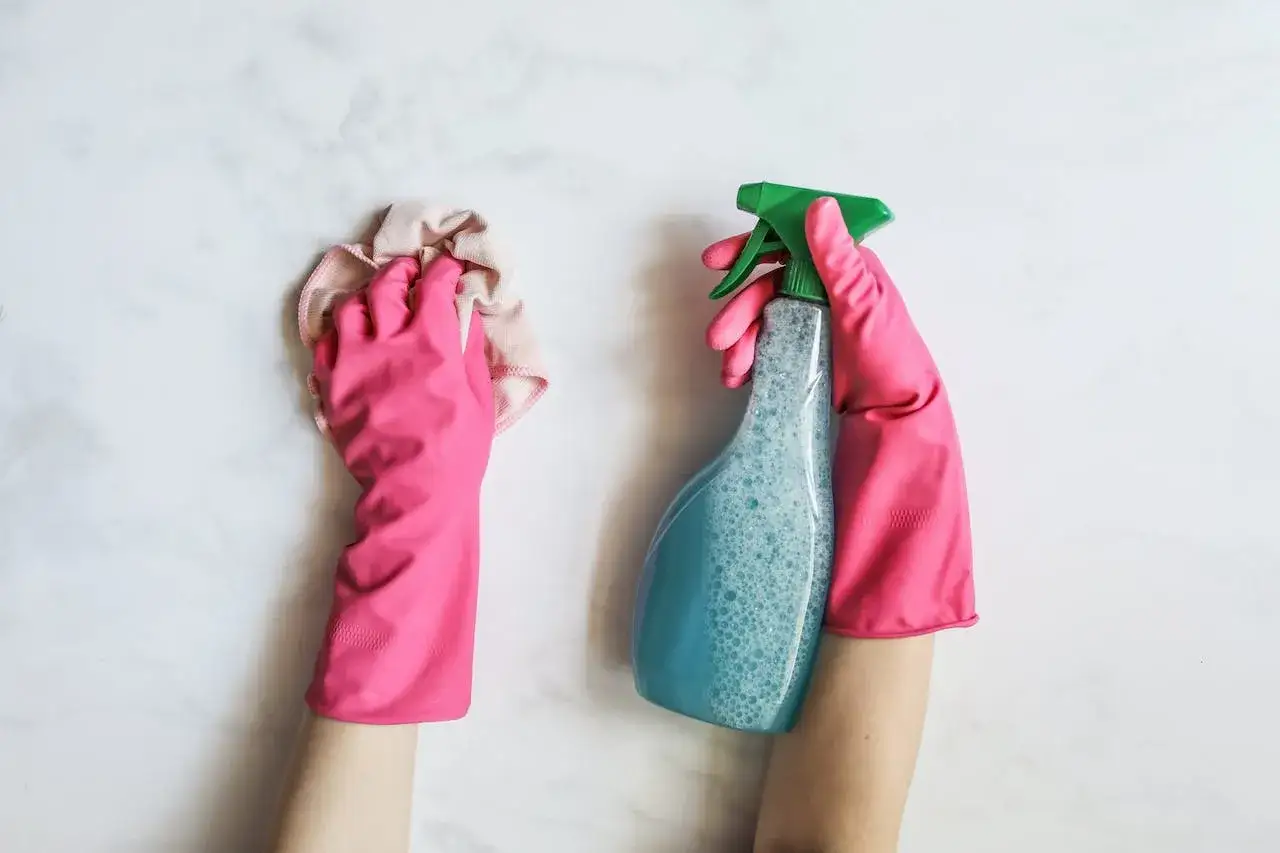 Manos con guantes rosas limpian manchas oscuras plato ducha con spray y pa&ntilde;o.