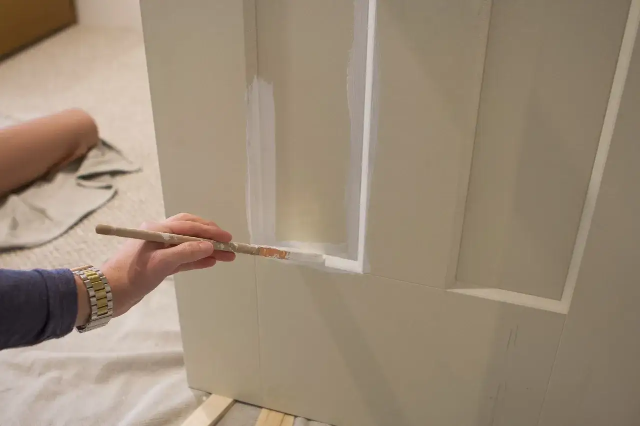 Mano pintando una puerta de madera con brocha blanca. Se aprecian detalles de la puerta y la mano con reloj.