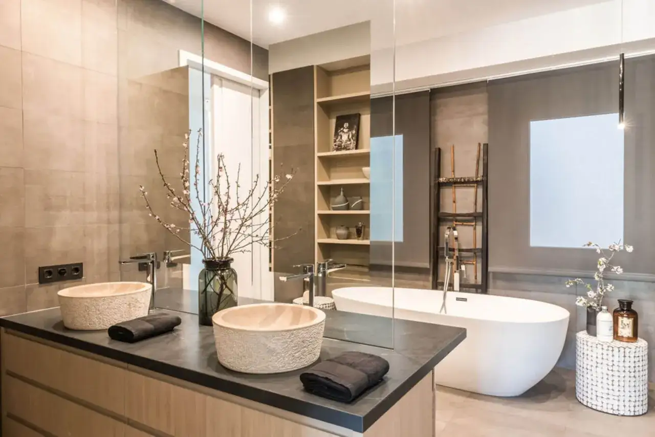 Ba&ntilde;o moderno con ba&ntilde;era exenta blanca, lavabos de piedra, encimera oscura y estanter&iacute;as. Averiguar cuanto cuesta poner una ba&ntilde;era es clave para este dise&ntilde;o.