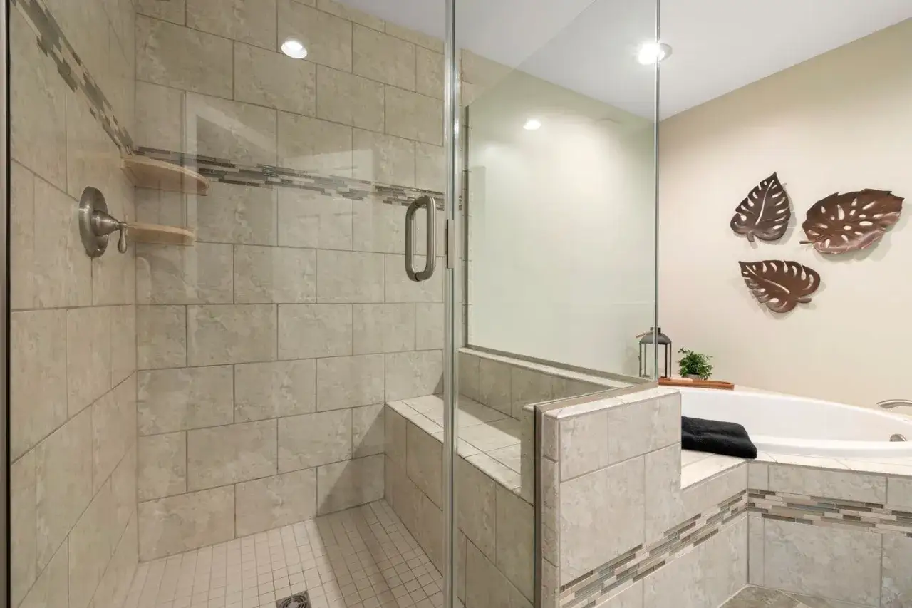 Duchas con murete y cristal, con banco integrado y azulejos de piedra. Bañera adyacente con decoración de hojas.