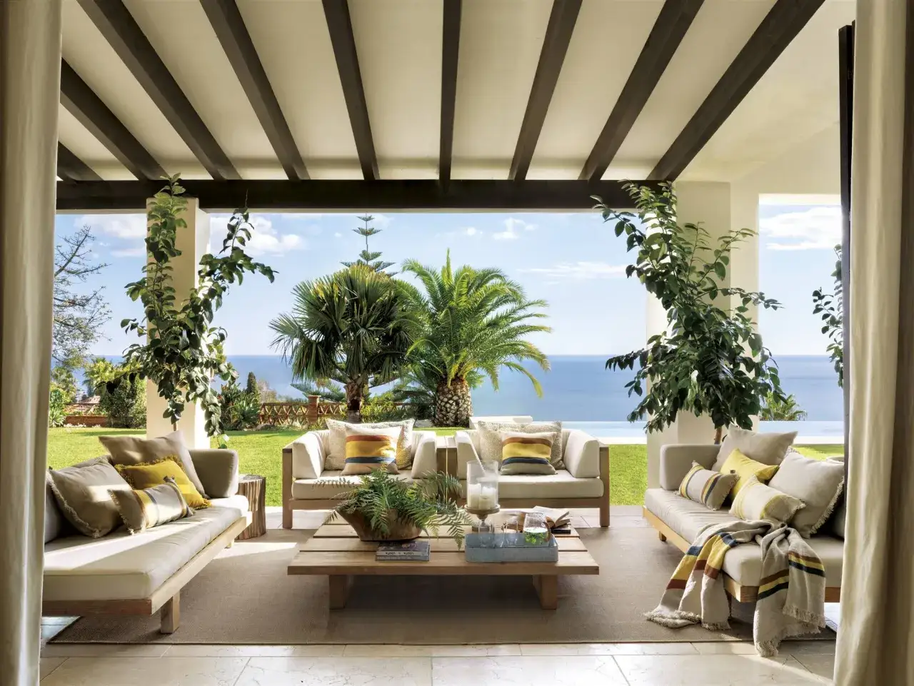 C&oacute;modos porches para patios con sof&aacute;s de madera, cojines y mantas, con vistas al mar y palmeras.