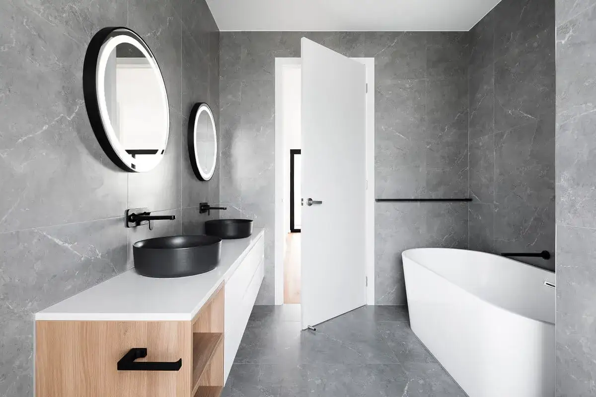 Ba&ntilde;o moderno con lavabos negros, ba&ntilde;era exenta y suelo radiante en ba&ntilde;os. Azulejos grises y detalles en madera.