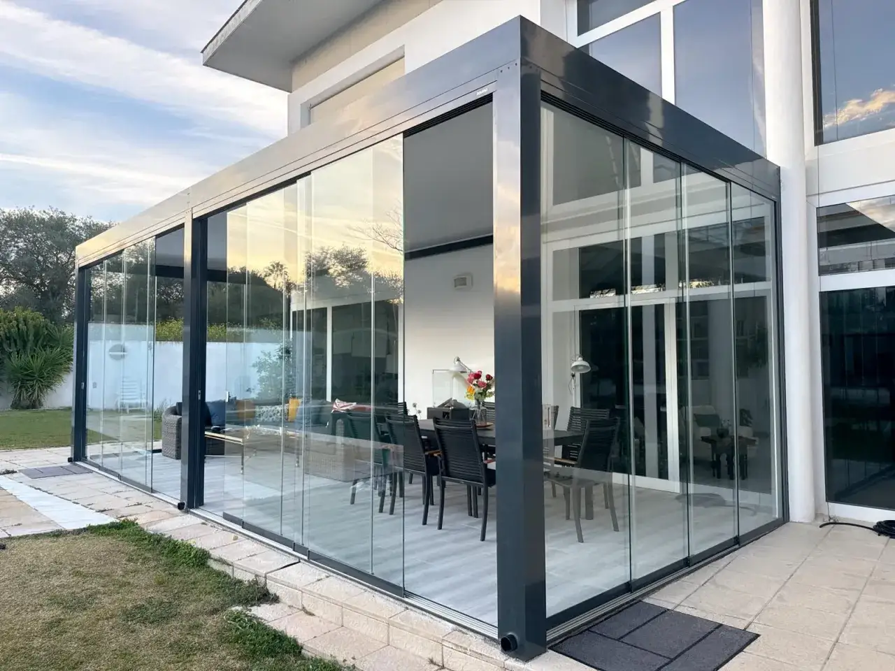 Moderno cerramiento abatible terraza con cristales que permiten disfrutar del exterior. Interior amueblado con mesa y sillas.